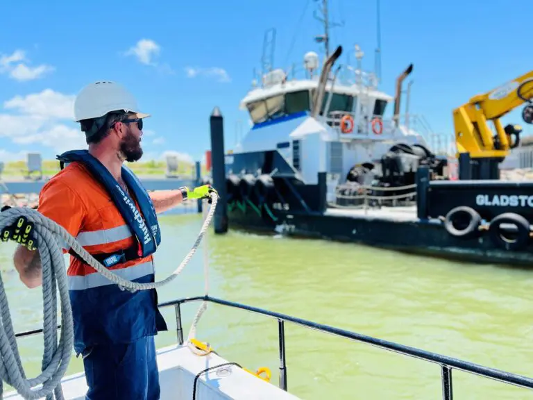 Worker on a boat deck pulling a mooring rope, wearing a safety helmet, high-visibility shirt and life jacket, with a tug boat in the background